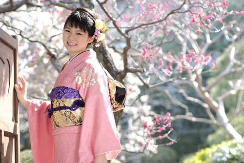 Japanese woman in a kimono posing near cherry blossom.
