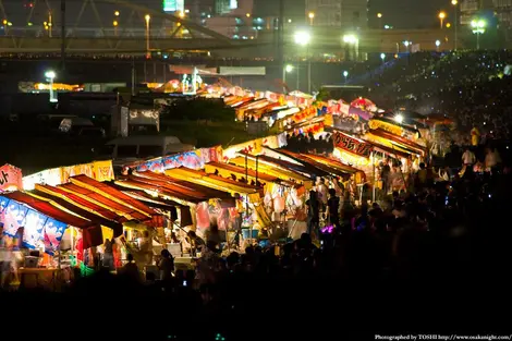 En las orillas del río Yodogawa, las multitudes luchan por obtener los mejores puestos para ver los fuegos artificiales. En las orillas del río Yodogawa, las multitudes luchan por obtener los mejores puestos para ver los fuegos artificiales.