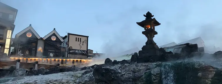 Stone lantern on top of a rock with steam coming from the ground and buildings in the background. 